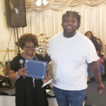 older woman holds a certificate while standing next to a young man in a white shirt, both smiling inside a room with musical instruments in the background.