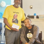 older man in a yellow staff shirt stands next to a smiling young woman in a basketball shirt sitting in a chair indoors.