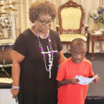 older woman stands beside a young boy holding a paper, with musical instruments and ornate chairs in the background.