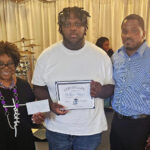 Three people posing indoors; the person in the center holds a certificate, while the woman on the left holds an envelope.