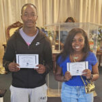 Two people standing indoors, smiling and holding certificates in their hands, with a curtain and flowers in the background.