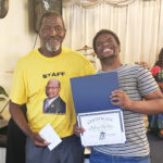 Two smiling men, one holding a certificate and a blue folder, standing indoors. The older man wears a yellow "staff" shirt and holds a card.