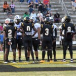 Football players from two teams meet on the field for the coin toss, with referees present and spectators seated in the stadium bleachers in the background.