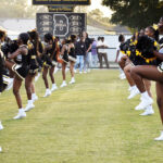 Cheerleaders in black and yellow uniforms perform a routine on a football field, forming two lines with raised arms and legs. A scoreboard is visible in the background.