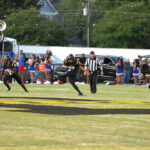 Football players running on a field during a game, with a referee and cheerleaders visible in the background, and a crowd watching from the sidelines.