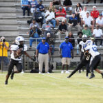 High school football game in progress, players in black uniforms on the field, coaches and spectators watching from the bleachers in the background.