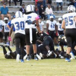 Football players in blue and white uniforms gather on the field around a pile-up during a game, with spectators seated in the bleachers in the background.