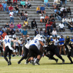 A high school football game in progress, with players in white and black uniforms competing while spectators watch from bleachers in the background.