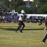 A football player in black and white uniform prepares to throw the ball during a game, with spectators and a marching band in the background.
