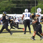 Football players in black and white uniforms are actively engaged during a game on a grassy field with spectators watching in the background.