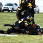 Football players in black uniforms tackle an opponent during a game on a grassy field, with cars and a white fence visible in the background.