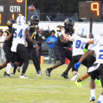 Football game action with players in black and white uniforms; quarterback prepares to throw the ball while defenders rush. Scoreboard and spectators visible in background.
