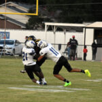Two football players in white uniforms tackling an opponent during a game on a grassy field, with spectators and a goalpost visible in the background.