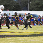 High school football game in action, players in black running with the ball, referee nearby, cheerleaders and spectators in the background.