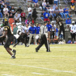 Football players in black and white uniforms compete on a grassy field, with a referee and crowd in bleachers in the background.