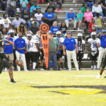 Two American football teams play on a field with coaches, players, and spectators visible in the background, along with an orange down marker on the sideline.