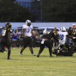 Football players in black and white uniforms compete on a grassy field during a game, with one player being tackled while others look on. Spectators are visible in the background.