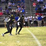 Two American football players in black uniforms running on a field during a game, with spectators and team members visible in the background.