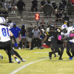Football players in black and white uniforms compete under stadium lights, with referees and spectators watching from the stands in the background.