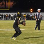 A football player in black uniform prepares to throw the ball during a night game, with a referee and spectators visible in the background.
