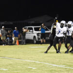 Football players in white uniforms celebrate on a field at night, with spectators, vehicles, and bright stadium lights in the background.