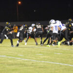 Football players in black and white uniforms compete under stadium lights at night, with players running and blocking on a grassy field.