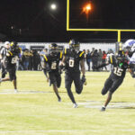 Football players in black uniforms running on a field at night, with a goalpost and crowd visible in the background.