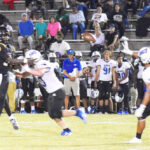 Football players in action during a game, with one player throwing the ball and opponents attempting to block, while coaches and spectators watch from the sidelines in the background.