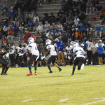 Football players in action on a field at night with a crowd of spectators in the stands. Some players wear white uniforms, others are in black.