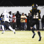 Football player in white uniform runs with ball while player in black, number 12, pursues; spectators watch from behind a fence on the sidelines at night.