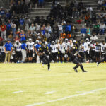 Two football players in black uniforms run on a field during a night game, with a crowd and opposing team in white on the sidelines.