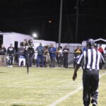 A football game at night shows a referee watching the field as players and a crowd stand along the sideline under bright lights.
