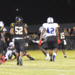 High school football players in black and white uniforms are in action on a lit field at night, with spectators and teammates watching from the sidelines.