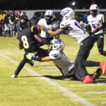 Football player in black jersey being tackled by two players in white uniforms during a nighttime game on a grassy field.
