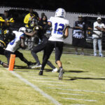 A football player in white is tackled near the end zone by players in black, while spectators watch from the sidelines at night.