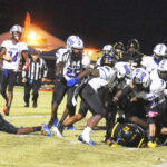 Football players in black and white uniforms pile up on the field during a nighttime game, with referees and bright lights in the background.