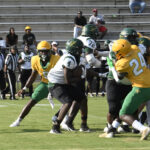 Football players in green and yellow uniforms compete on a field, with action occurring at the line of scrimmage. Spectators and referees watch from the background.