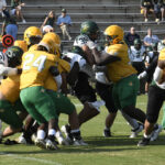 Football players in yellow and green uniforms tackle opponents during a game on a grassy field, with spectators watching from bleachers in the background.