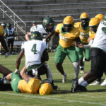 Football players in green and yellow uniforms tackle an opponent in white and green during a game on a grassy field. Bleachers are visible in the background.