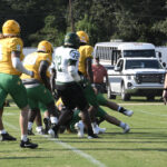 Football players in yellow and green uniforms compete on a grassy field, while a white vehicle and trees are visible in the background.