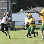 A football quarterback in a white jersey throws a pass as players in yellow and green attempt to block during a daytime game on a grassy field.
