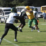 Football players in yellow and green uniforms compete on a grassy field, with school buses and white vans parked in the background.