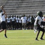 Football players practicing on a field, with one preparing to throw the ball and another ready to catch, while teammates watch from the sidelines in bleachers.
