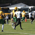 High school football players in green, yellow, and white uniforms run on a grassy field during a game, with school buses and a building in the background.