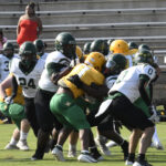 Football players in green, yellow, and white uniforms are engaged in a play on the field, with empty bleachers and a woman watching in the background.