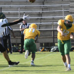 High school football game action, quarterback in yellow and green throws a pass while a defender in black and white attempts to block. Bleachers and referee in background.