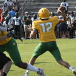 Football player in yellow jersey number 12 prepares to throw a pass during a game, with opposing team and spectators visible in the background.