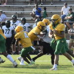High school football players in yellow and green uniforms compete on the field, while players in white uniforms and spectators watch from the bleachers in the background.