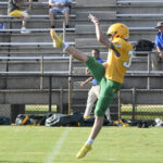 A football player in yellow and green uniform punts the ball during a game; empty bleachers and a few spectators are in the background.