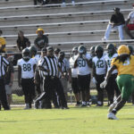 A football game in progress, with referees and players on the field and team members standing on the sideline near empty bleachers.
